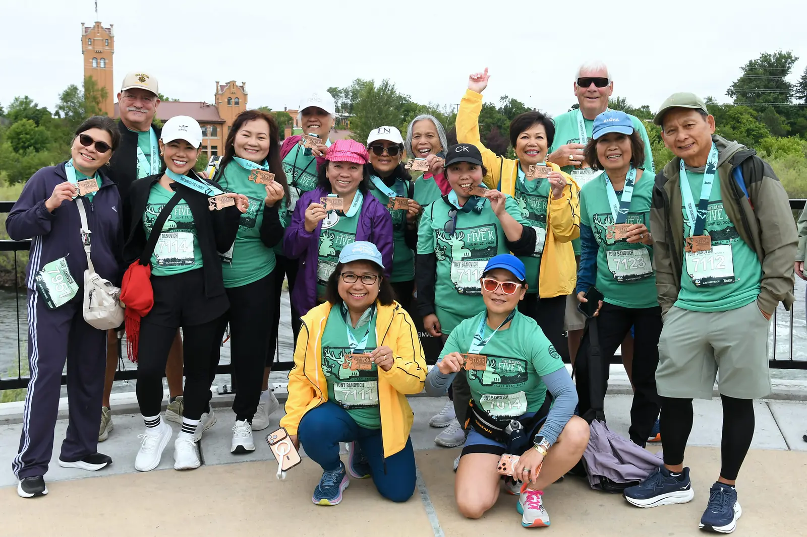 A large group of people in matching event shirts pose for a photo outdoors while wearing medals.