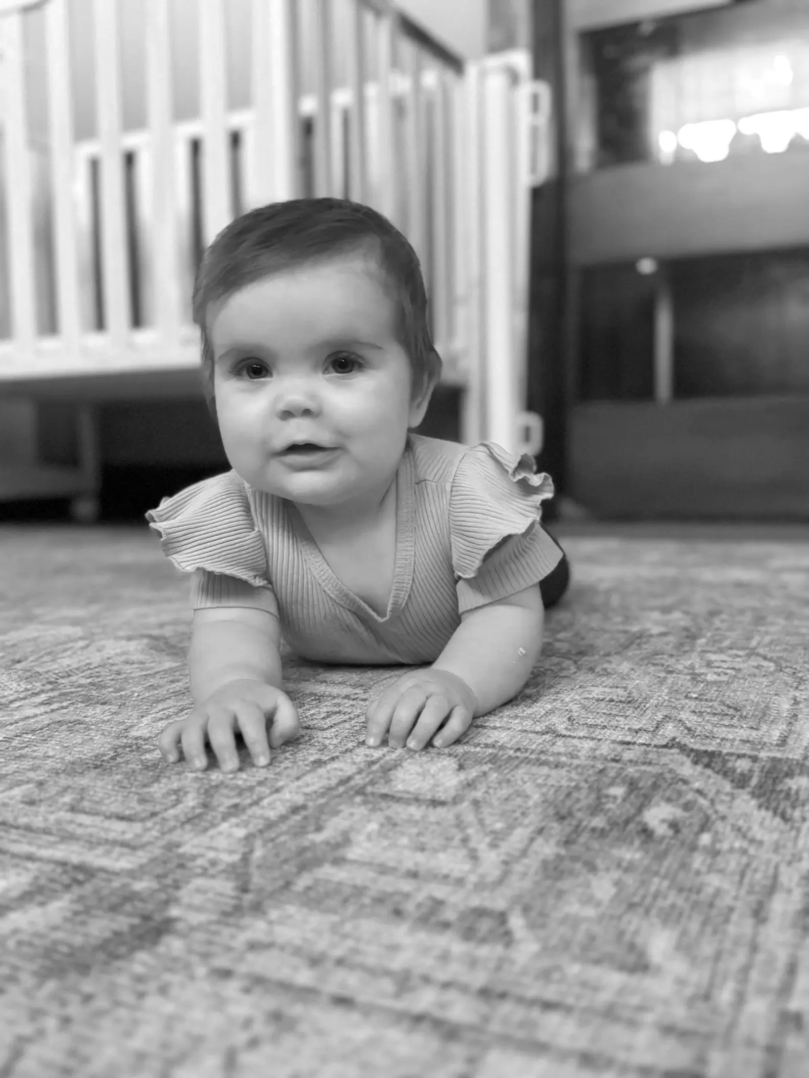 A baby lies on their stomach on a patterned rug in front of a crib.