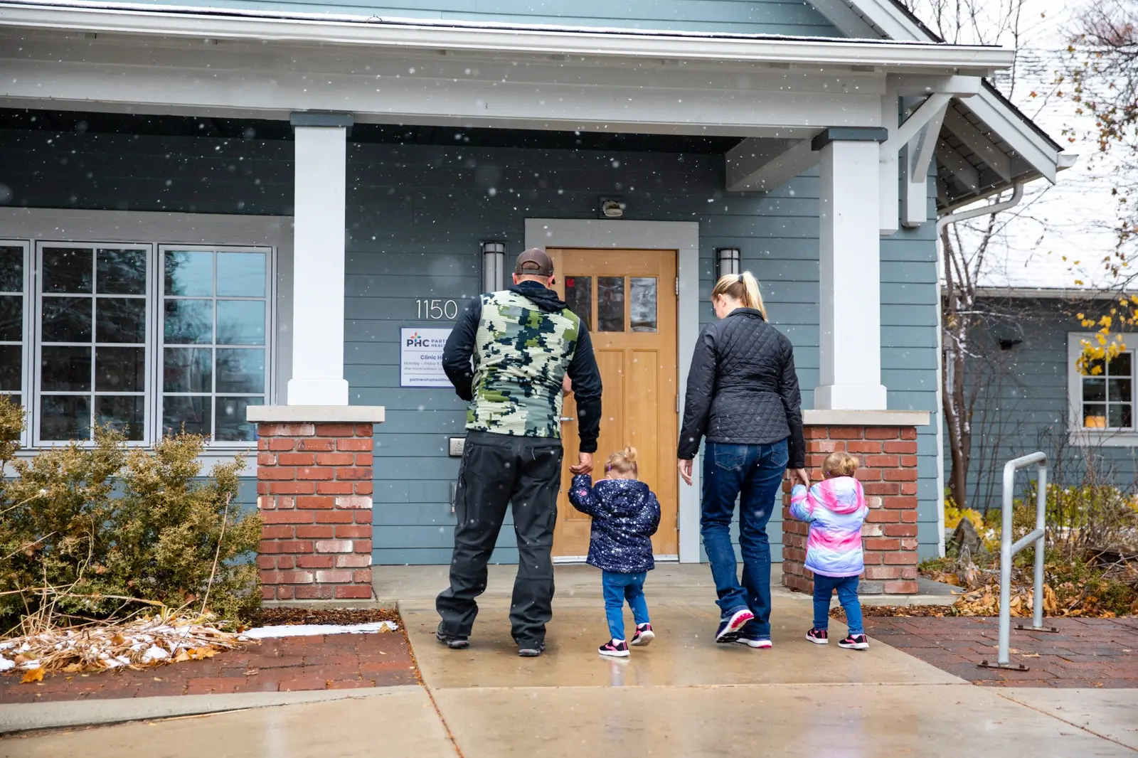 A family of four walks hand-in-hand toward the entrance of a building during a light snowfall.