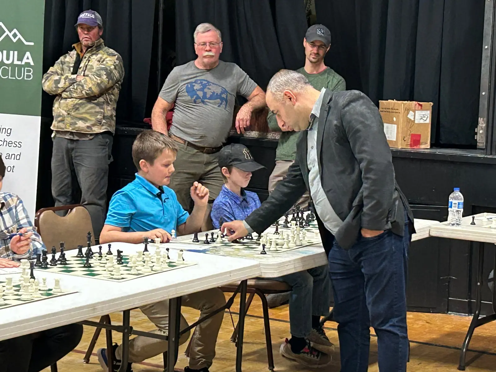 A man observes a boy playing a chess match as other people watch in the background.