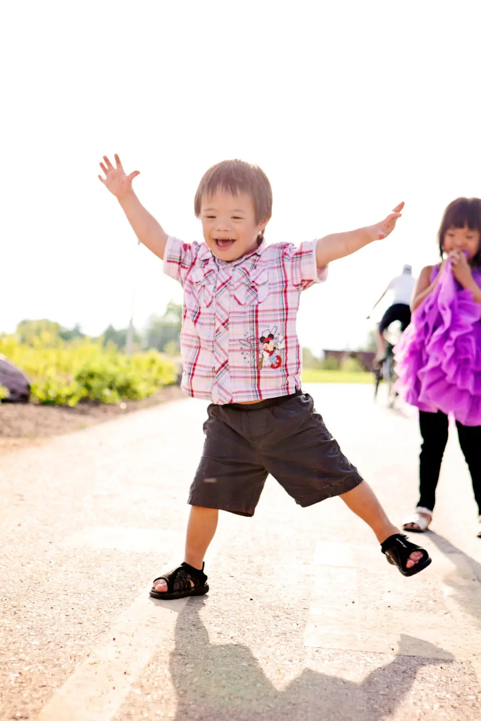 A young boy jumps excitedly on a paved path while a girl in a purple dress watches nearby.