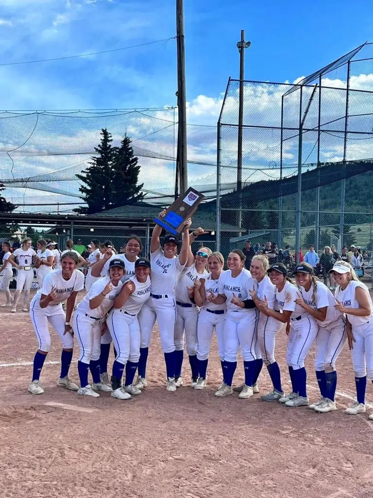 A softball team in uniform poses on a dirt field while holding a trophy.