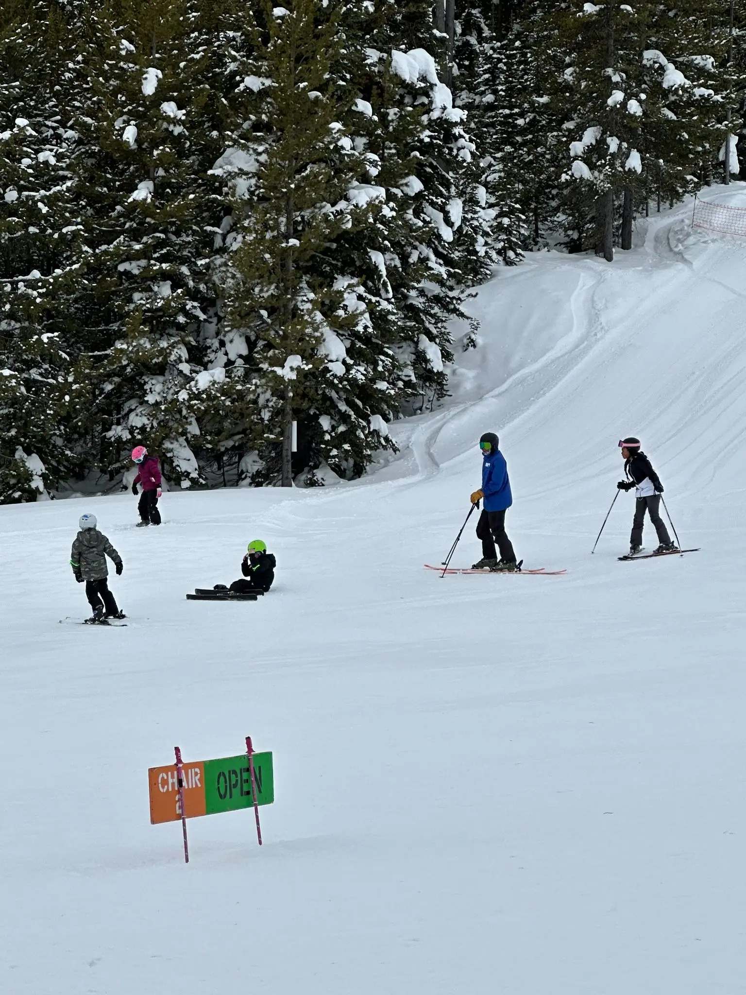 A group of people ski down a snow-covered slope near a sign indicating a ski lift is open.
