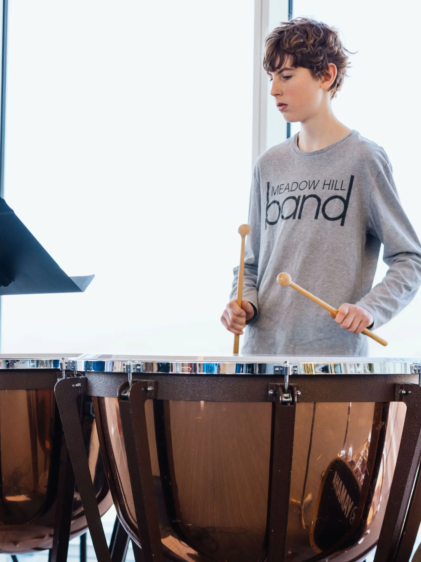 A young person wearing a band shirt plays the timpani with mallets.
