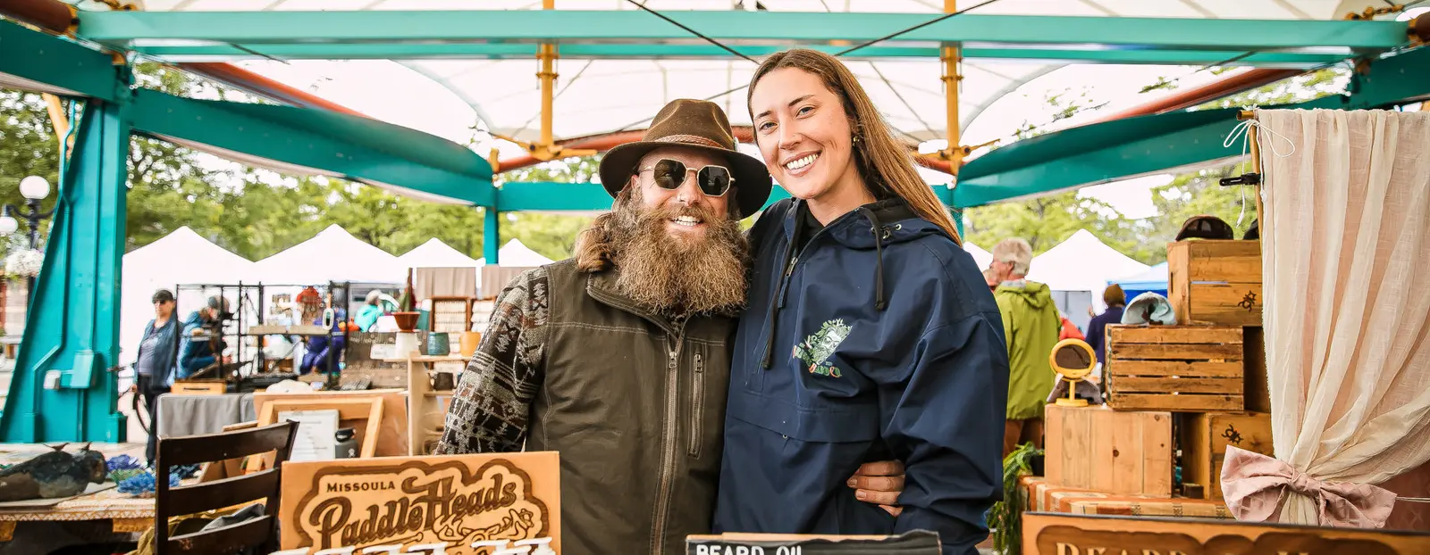 A man and a woman pose for a photo at an outdoor market stall.