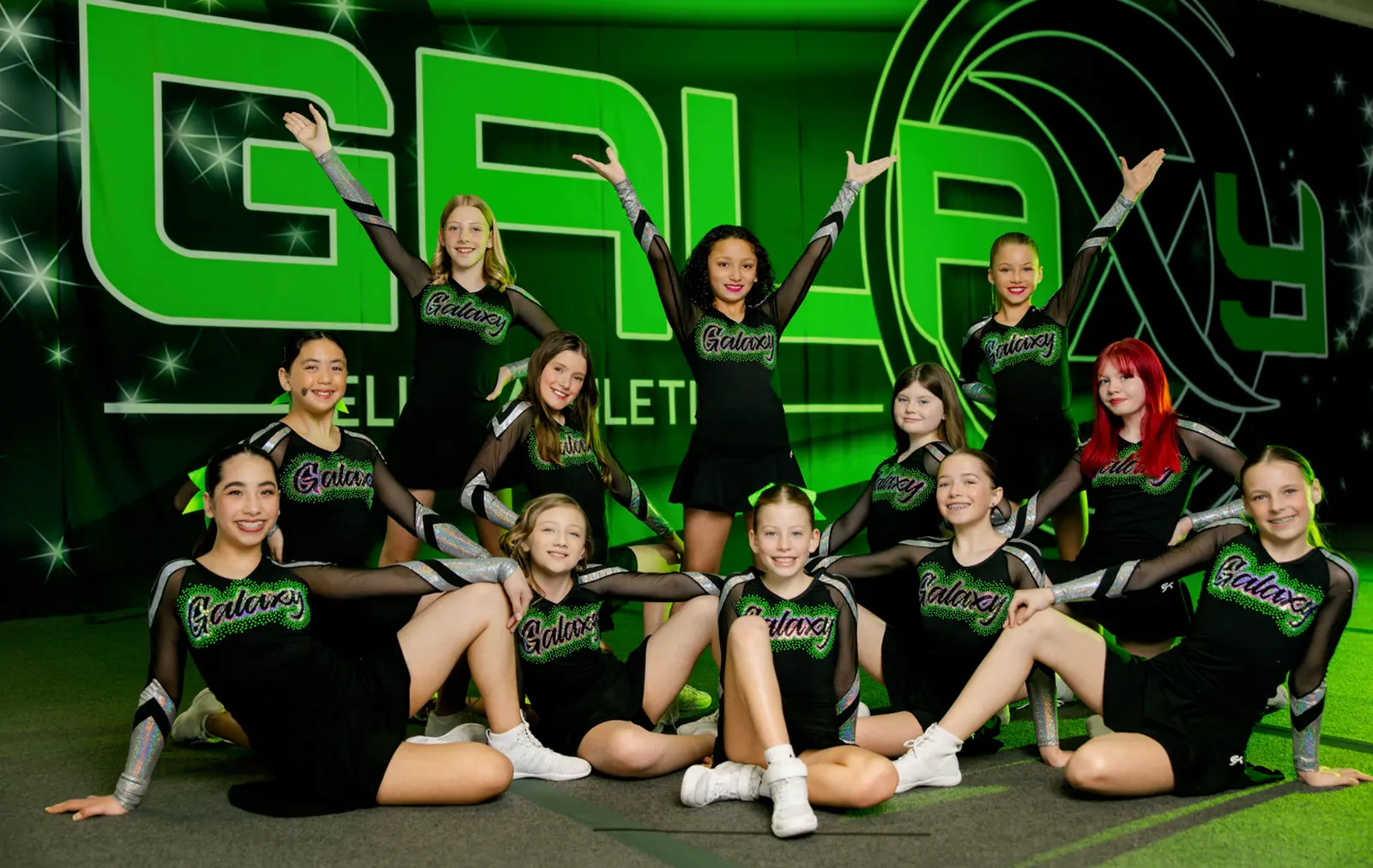 A group of young cheerleaders in uniform poses in front of a green and black branded backdrop.