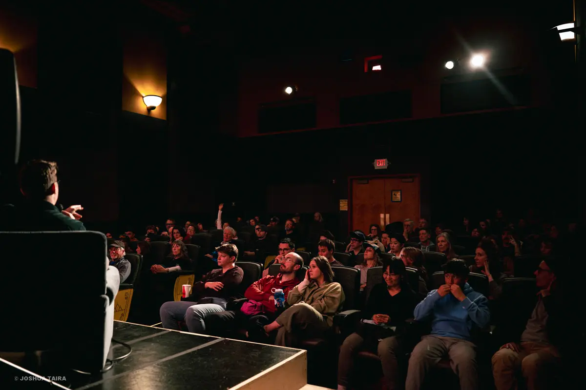 An audience sits in a dark theater watching a presenter on stage.