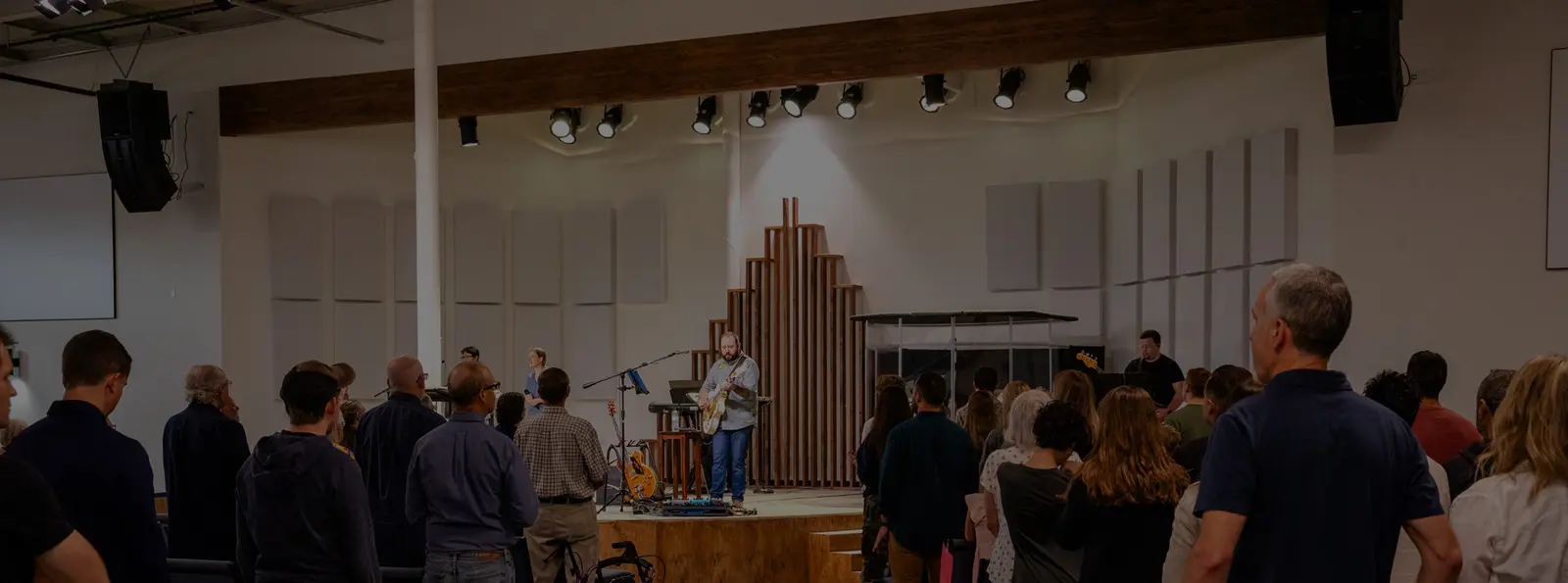A group of people stands in a large room while a musician plays a guitar on a stage.