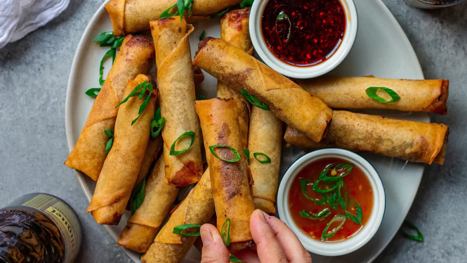 A hand holds one of several crispy fried spring rolls garnished with green onions on a plate with two dipping sauces.