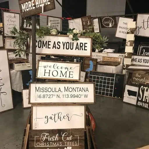 Display of decorative signs with welcoming messages, location details, and seasonal themes at a store or craft fair.