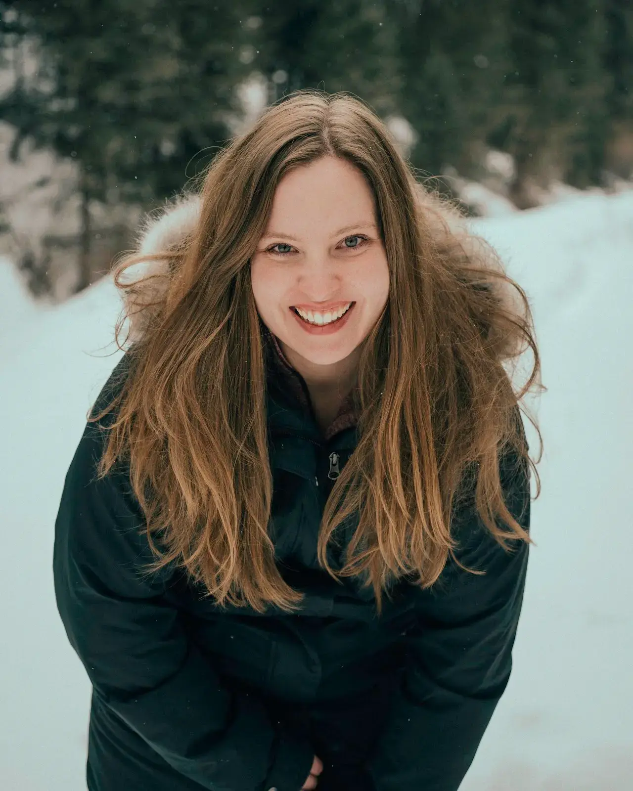 A smiling woman in a winter jacket stands outside in the snow.