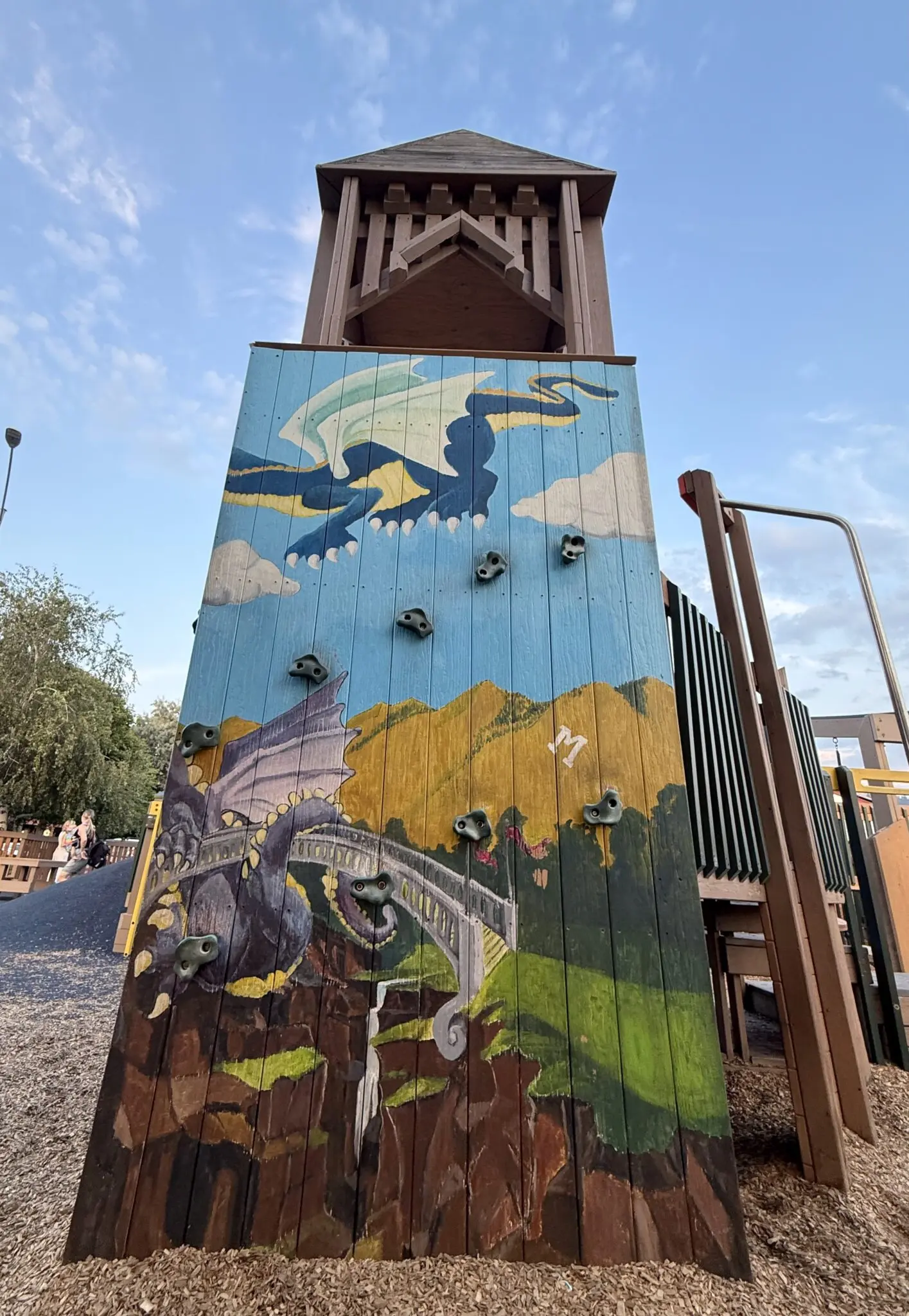 A wooden climbing wall at a playground features a painted scene of a dragon above a bridge.
