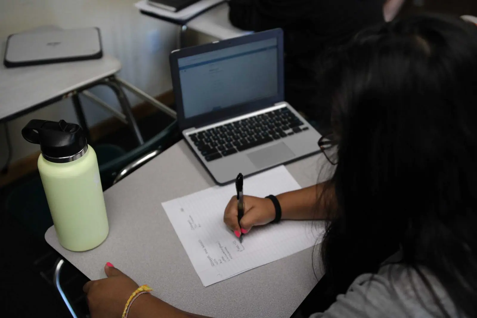 A student works at a desk with a laptop and a notebook in a classroom setting.