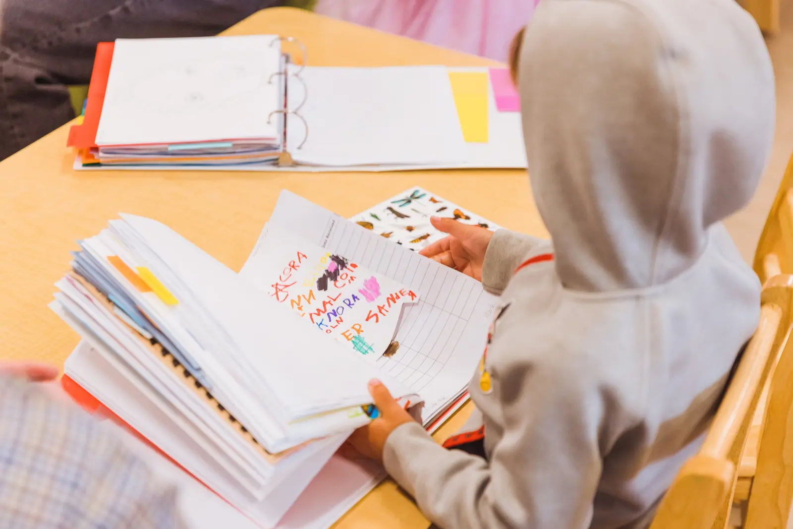 A child sits at a table and looks through a binder filled with papers and drawings.