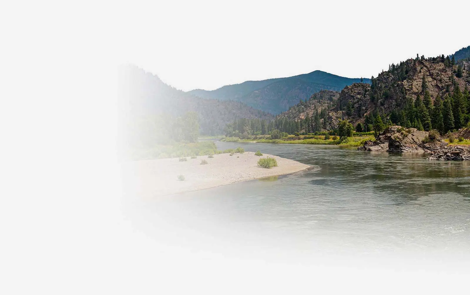 A wide river flows past a rocky shoreline under a mountain range.