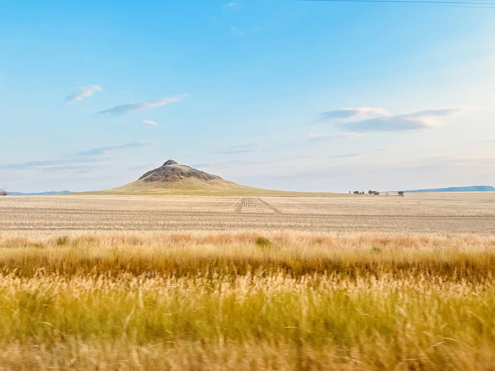 A lone hill rises from a vast, dry, harvested field under a clear blue sky.