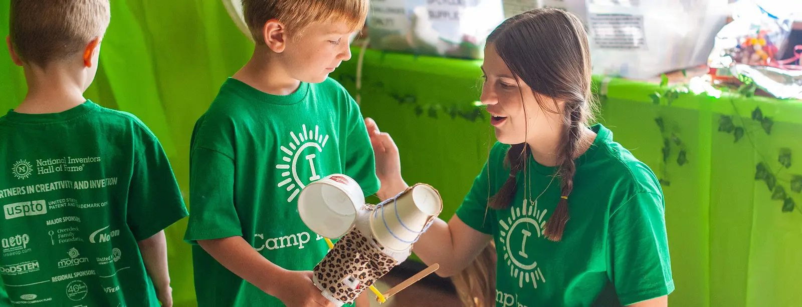 A Camp Invention counselor encourages a young camper to keep improving their invention prototype with recyclable materials