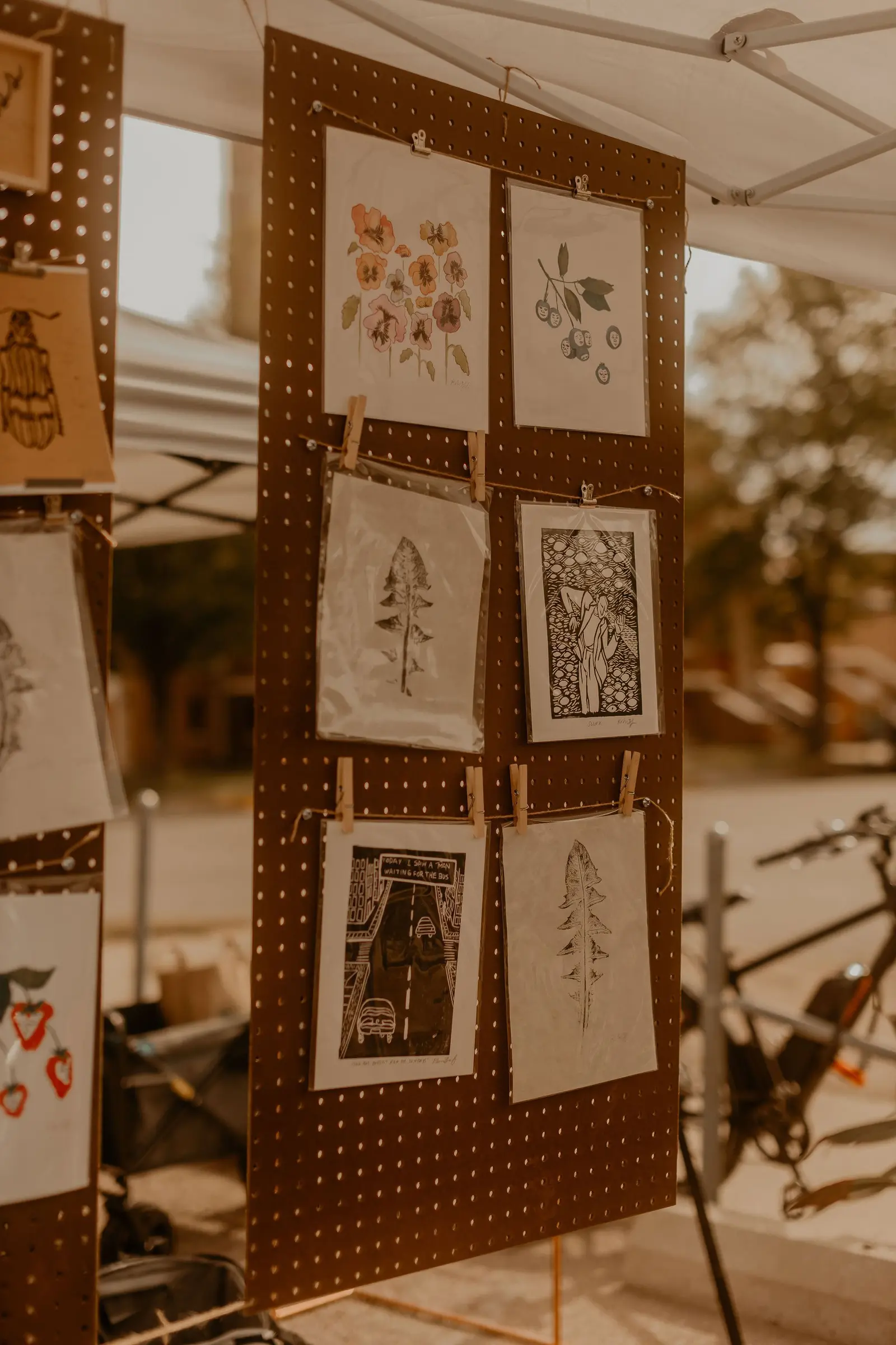 Display of various artwork hanging on a brown pegboard at an outdoor market, with some images of flowers, cherries, and abstract or stylized figures.