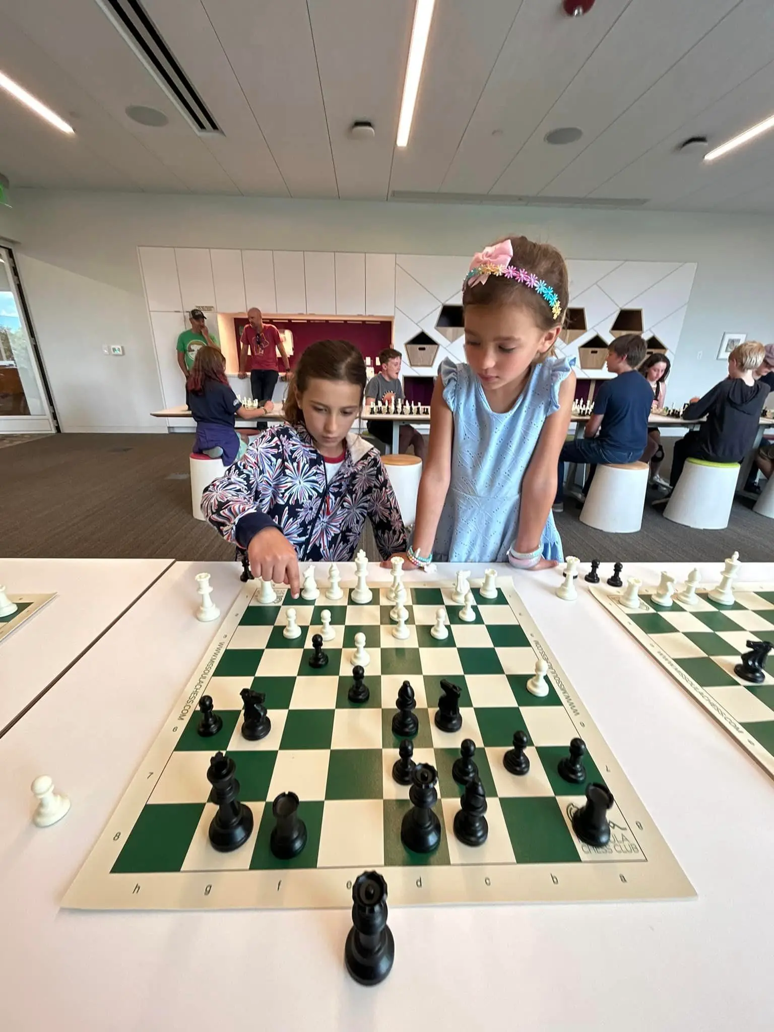 Two young girls play a game of chess together while others watch in the background.
