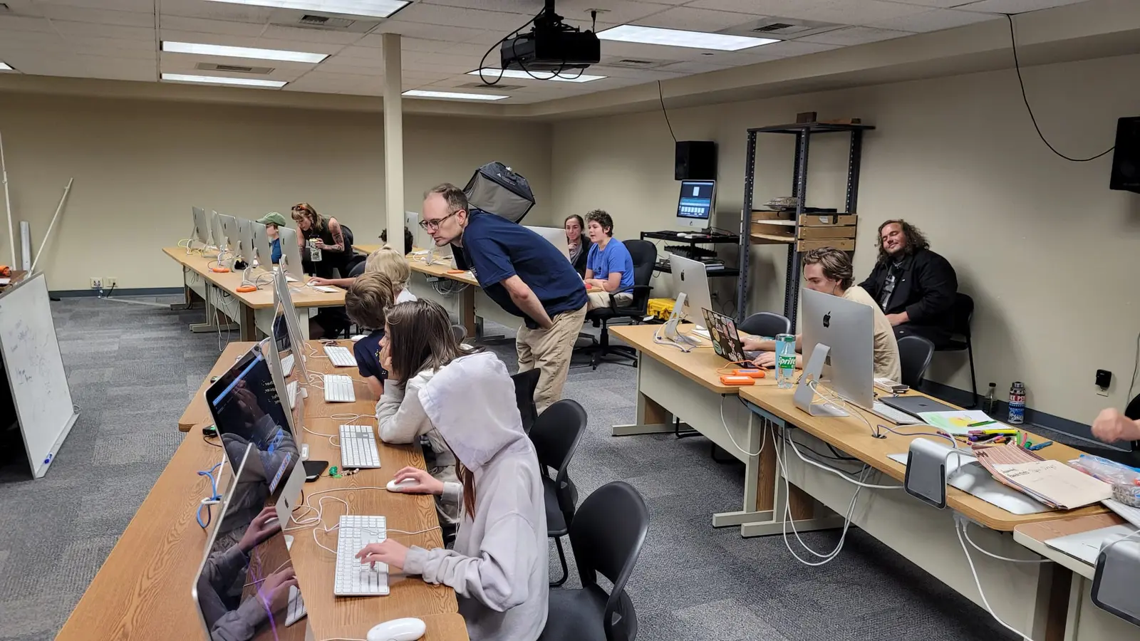 An instructor leans over to assist a student in a classroom setting with several rows of computers.