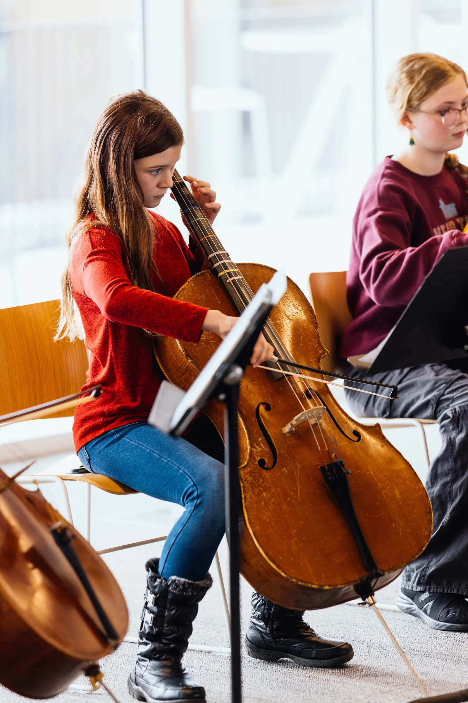 A young girl plays the cello in a room with other students.