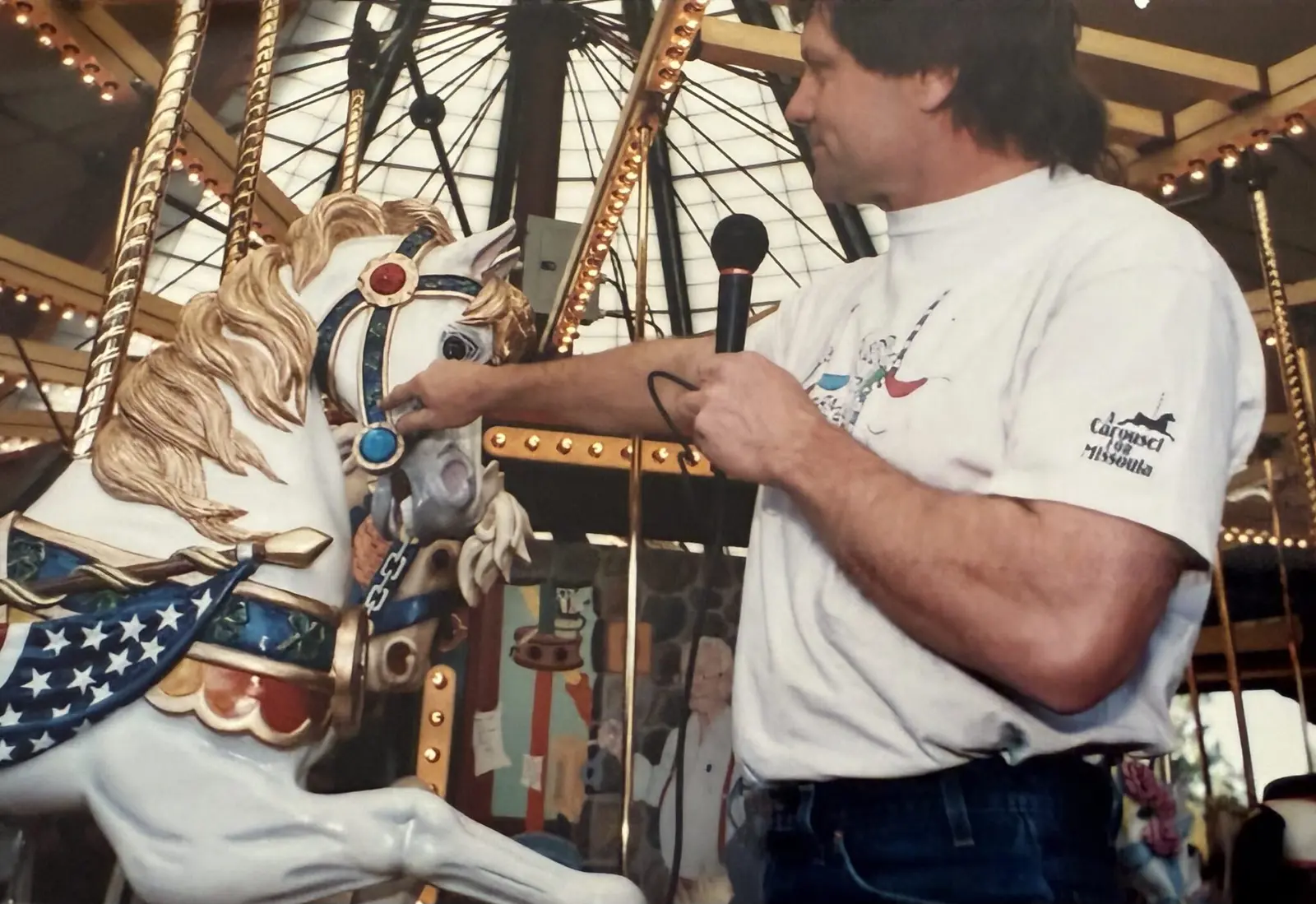 A man holds a microphone while pointing to a decorative carousel horse.