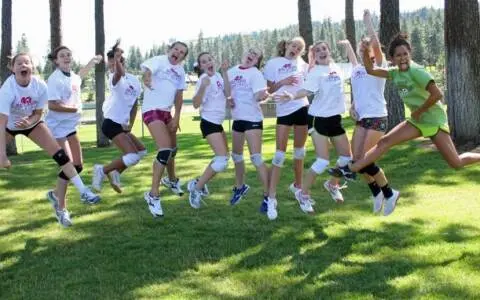A group of young women wearing matching t-shirts jump together in a grassy area with trees in the background.
