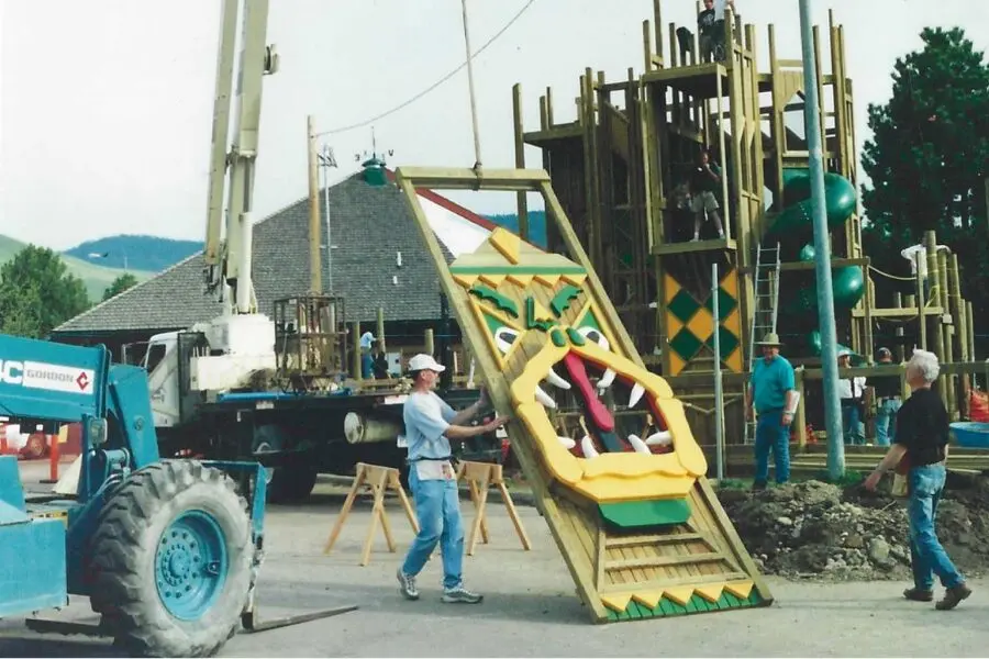 Construction workers use a crane to install a large decorative panel onto a playground structure.
