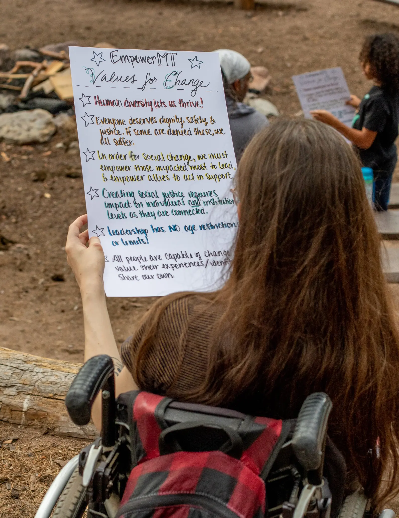 A person in a wheelchair holds a handwritten sign outlining values for social change.