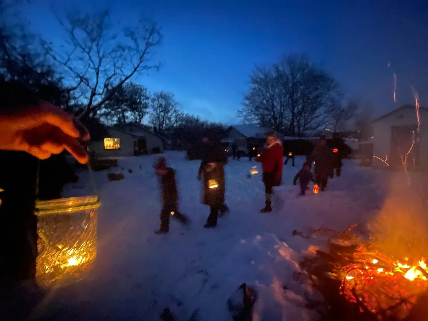 A group of people carries lanterns through a snowy area at dusk near a bonfire.