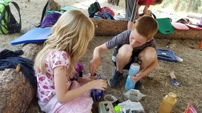 Two children sit on the ground outdoors, engaged in an activity with small items and containers.