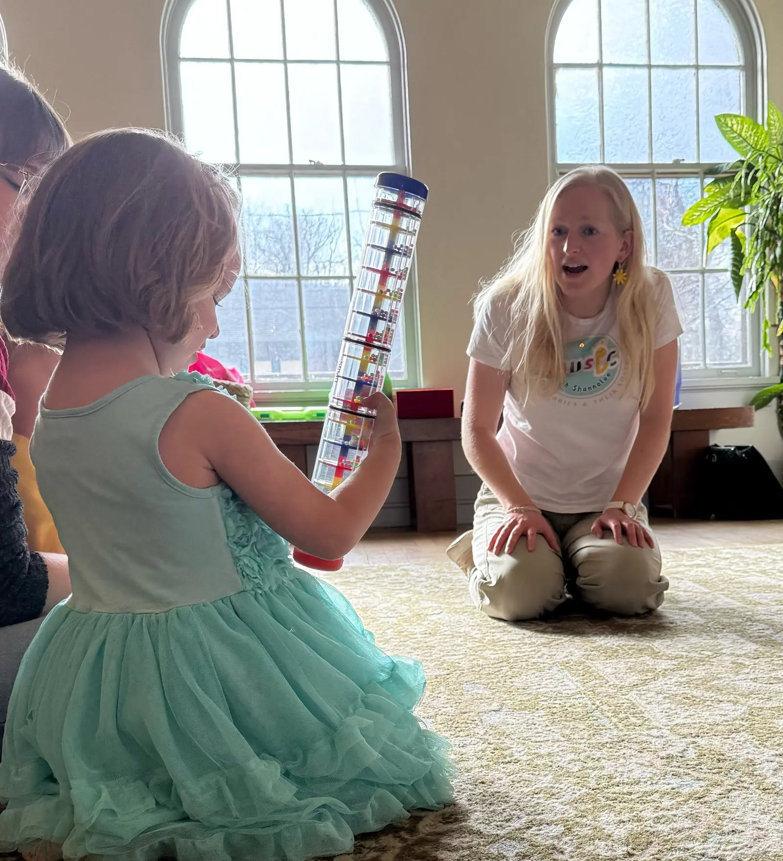 An adult sits on a rug while a young child holds a long, multi-colored sensory tube.