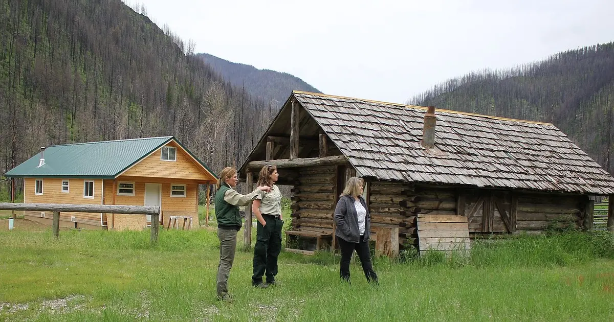 Three women stand in a field in front of two cabins with a mountain background showing signs of a fire.