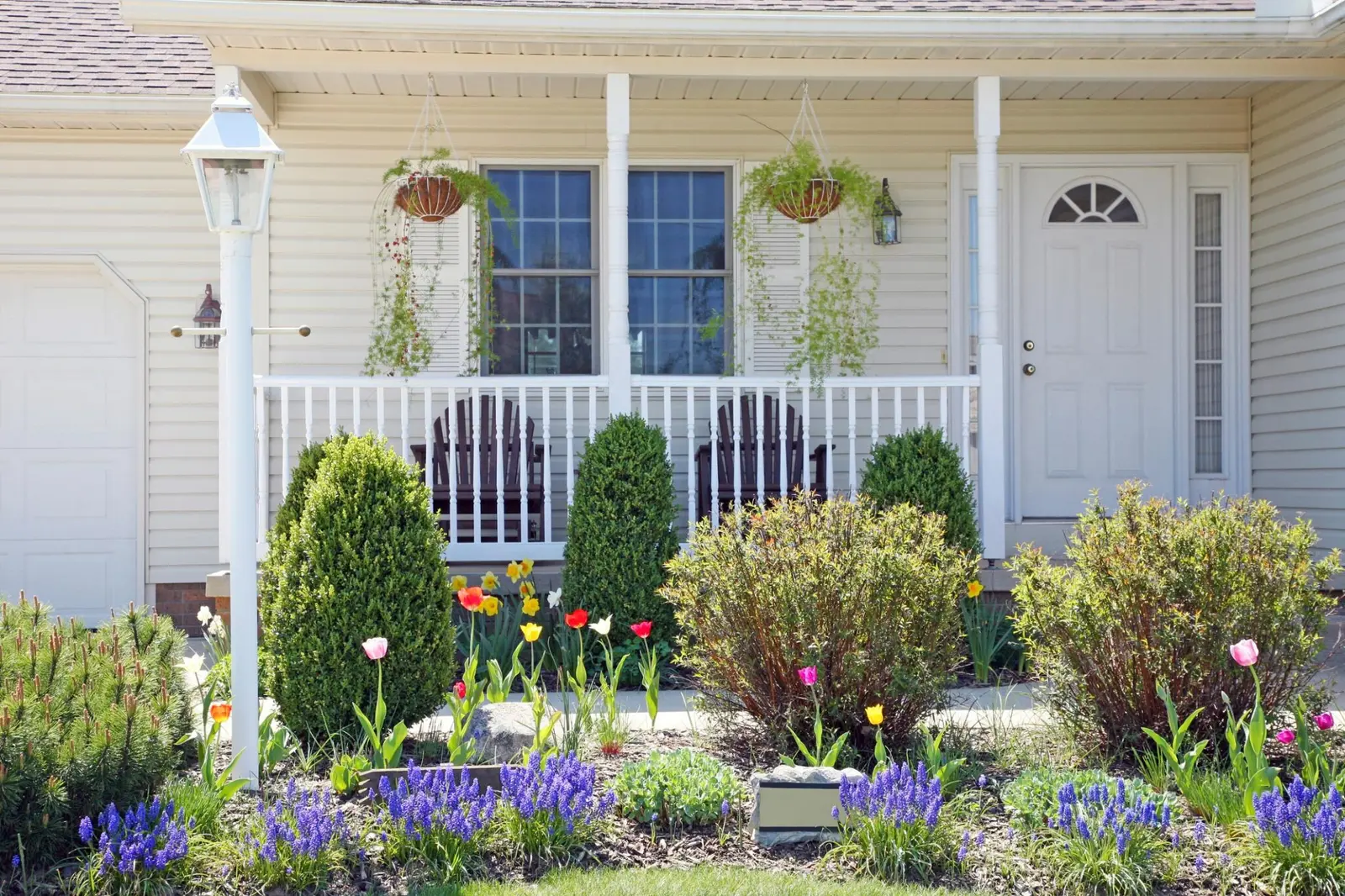 A well-maintained house front features a porch with hanging plants, Adirondack chairs, and a garden bed filled with blooming flowers and shrubs.