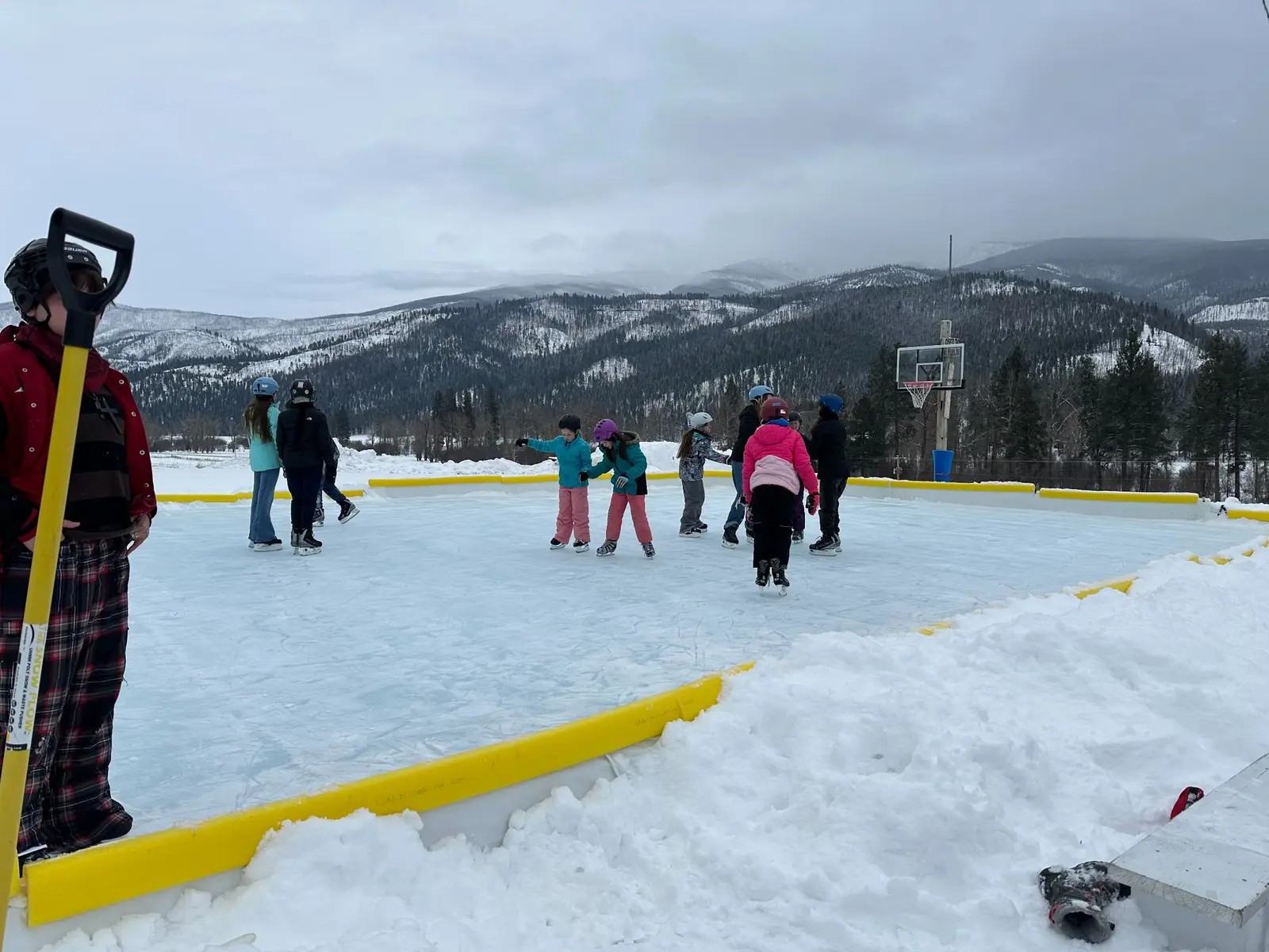 A group of children skates on an outdoor ice rink with mountains in the background.