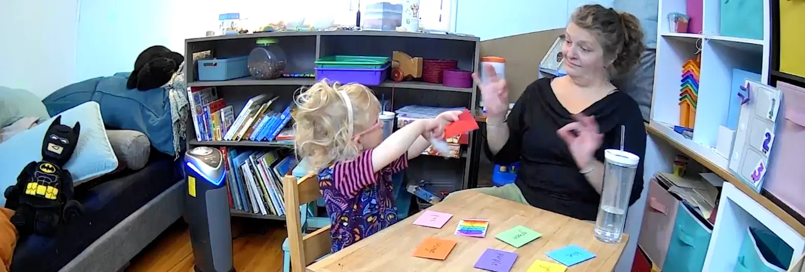 A teacher interacts with a young child while sitting at a table with colored cards.