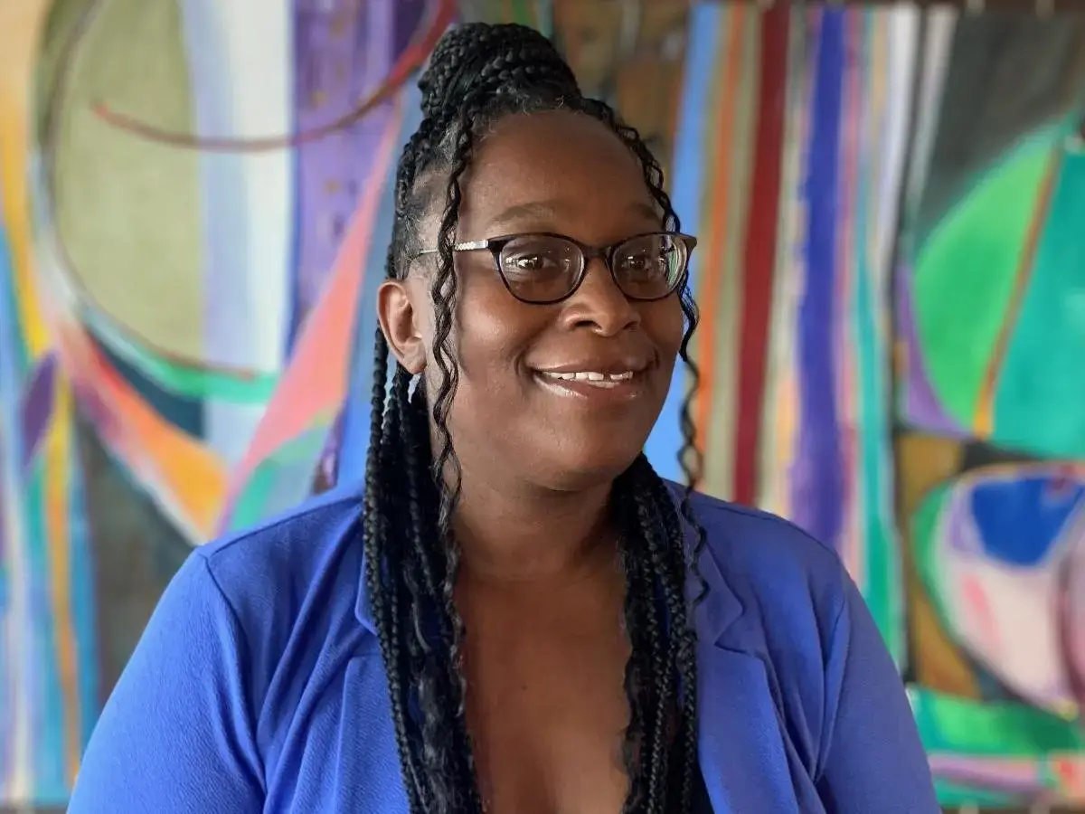 A smiling woman with braids wears glasses and a blue blazer in front of a colorful abstract art piece.