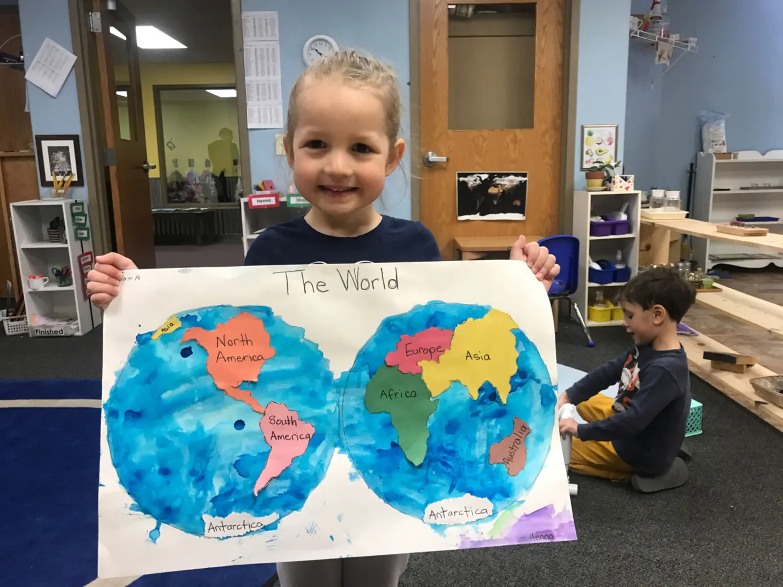 A young girl holds her handmade world map project in a classroom while another child works nearby.