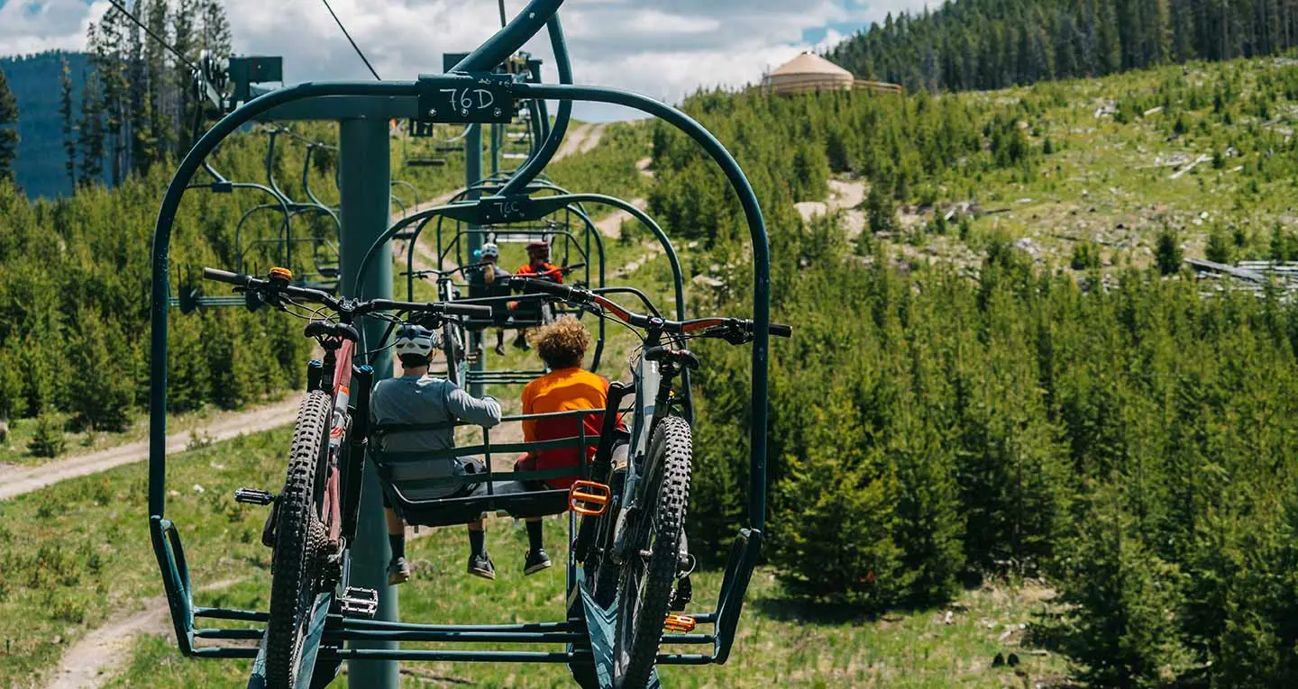 People ride a chairlift while carrying mountain bikes over a wooded hillside.