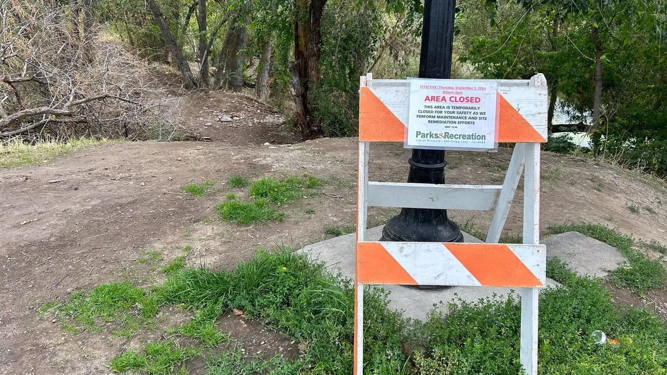 A temporary closure sign is attached to a barricade standing on a dirt path in a park area.
