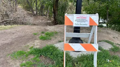 A temporary closure sign is attached to a barricade standing on a dirt path in a park area.