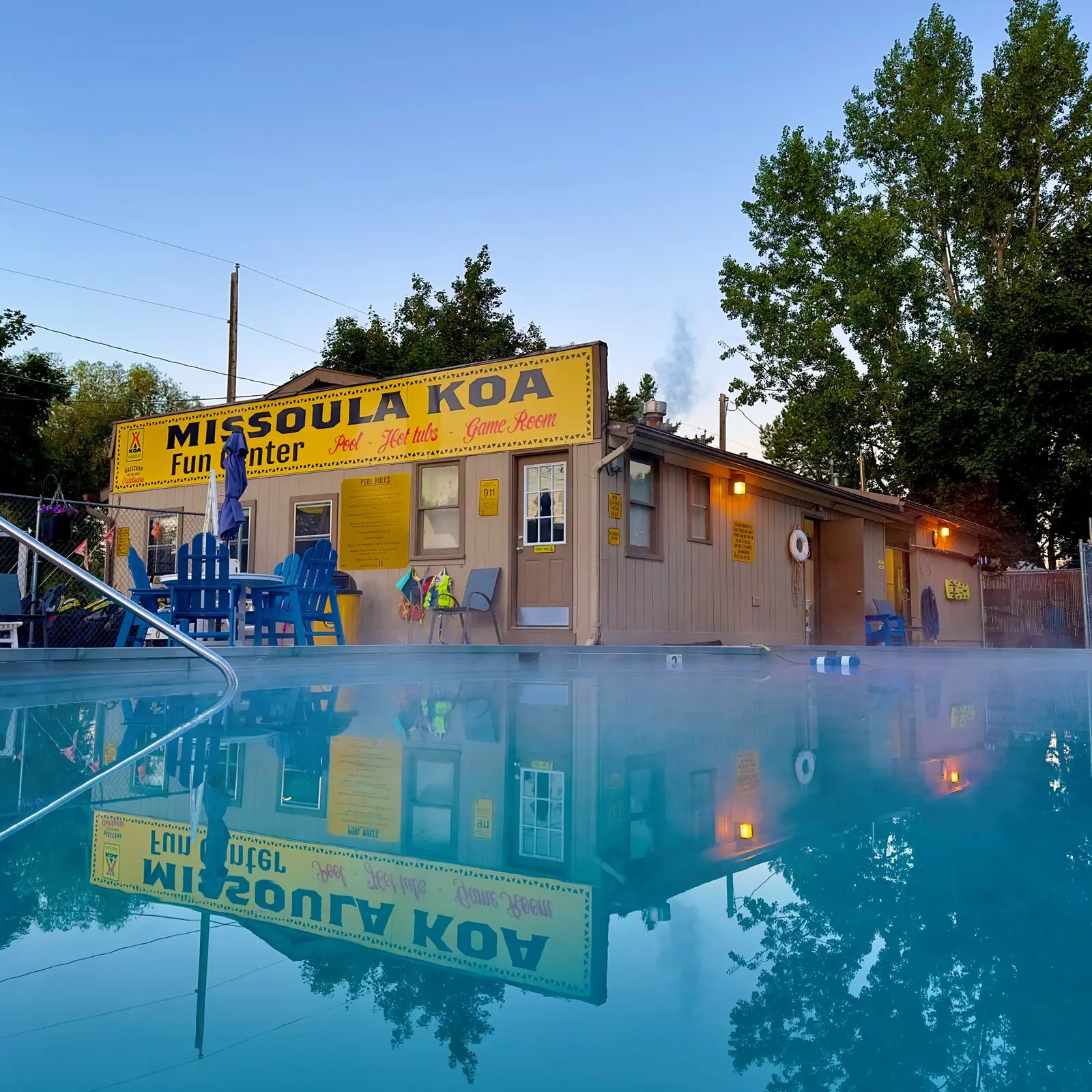 A building with a sign and pool deck is reflected in the water of a pool.