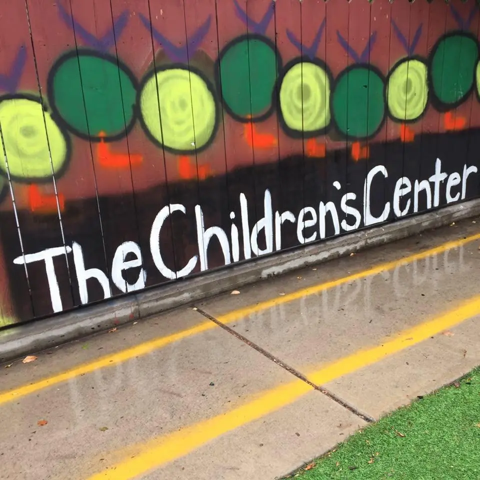 A wooden fence is decorated with a mural featuring green circles and the text "The Children's Center."