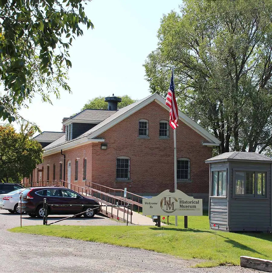 A brick historical museum building features a parking lot in front and a welcome sign.
