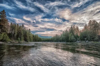 Photo from missoulacurrent.com A river flows through a forest under a cloudy sky.
