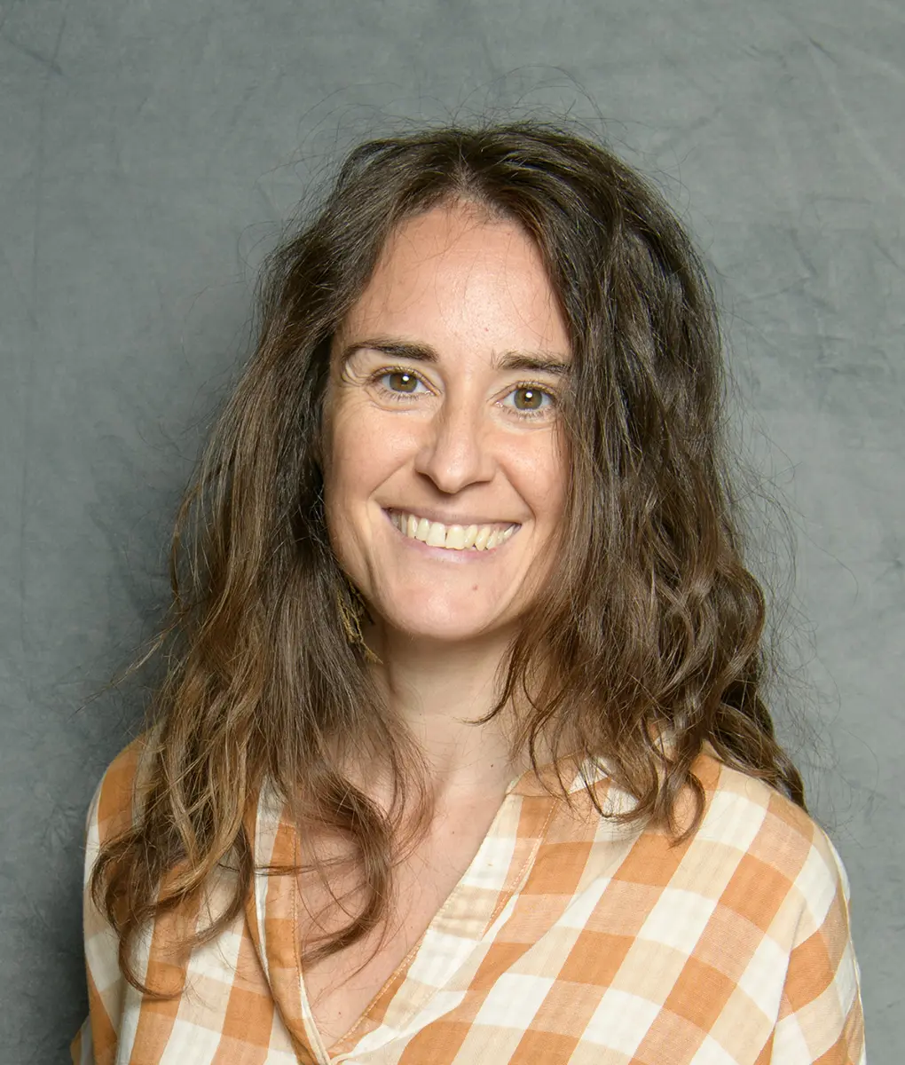 A smiling woman with long wavy hair is wearing a checkered shirt.