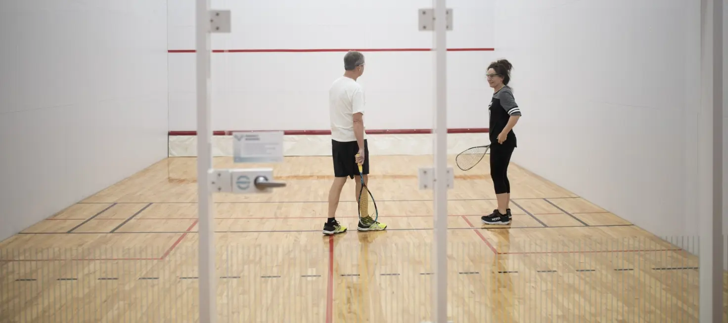 Two adults stand in a squash court holding rackets.