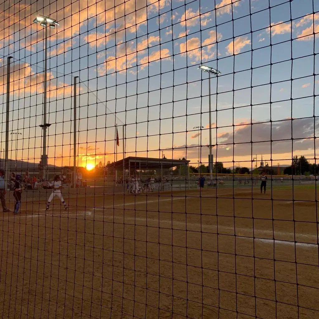 A youth baseball game takes place on a field during a sunset viewed through a safety net.