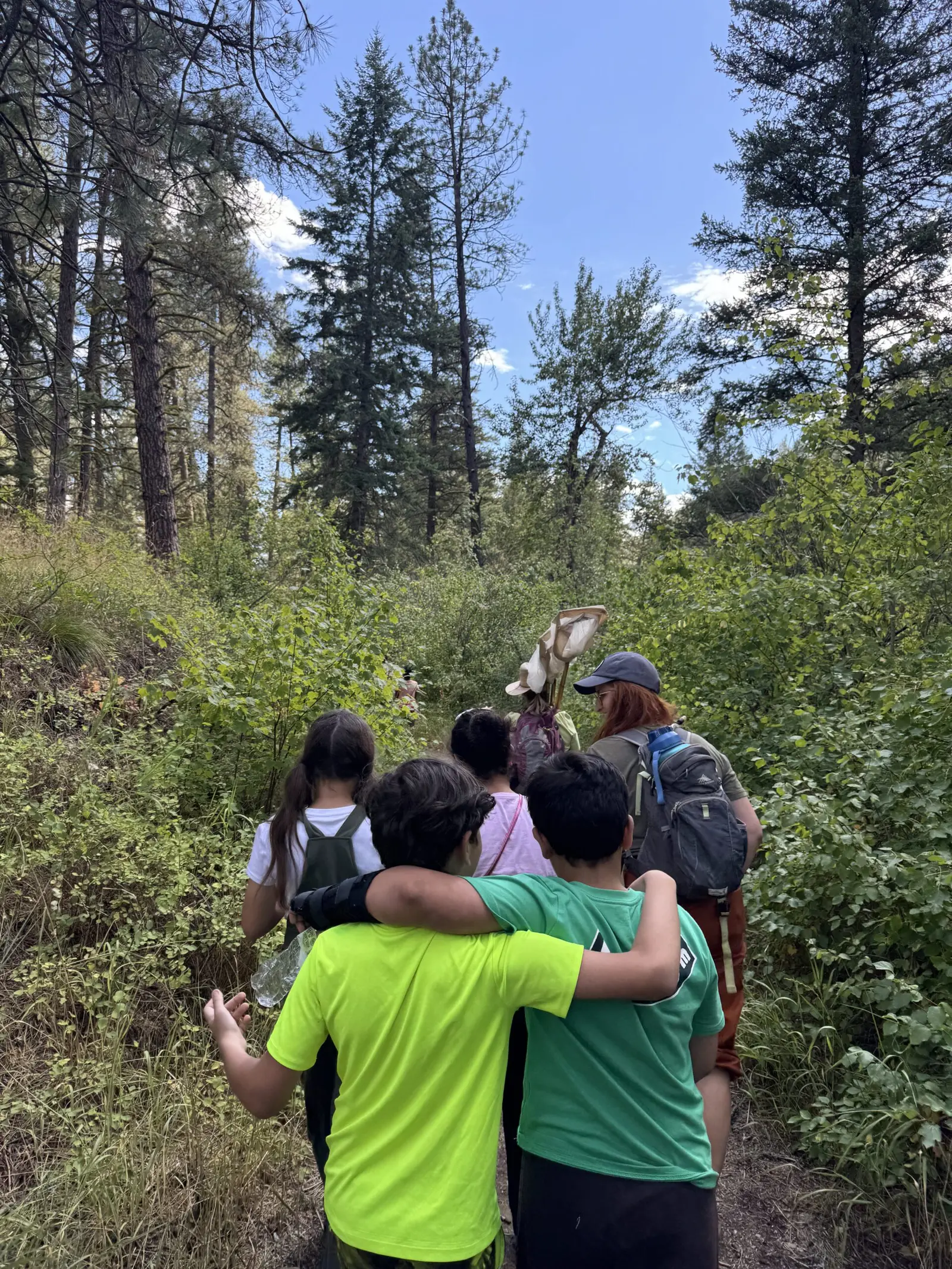 A group of children and an instructor are walking along a forested trail.