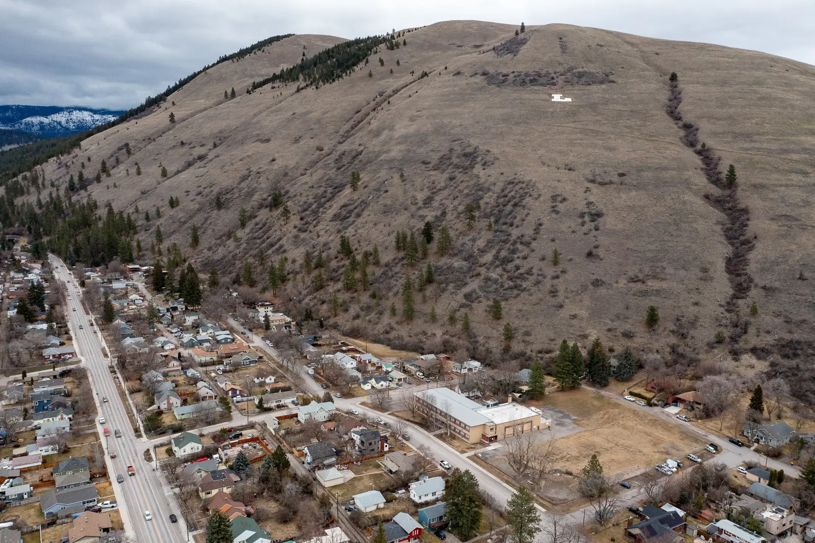 An aerial view shows a small residential town nestled at the base of a large, grass-covered hill featuring a white letter emblem.